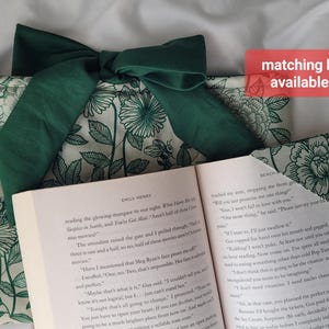 May include: A book sleeve and matching bookmark with a floral pattern in green and white. The book sleeve has a large green bow. The open book displays text. The text "matching bookmark available in shop!" is in a red box.
