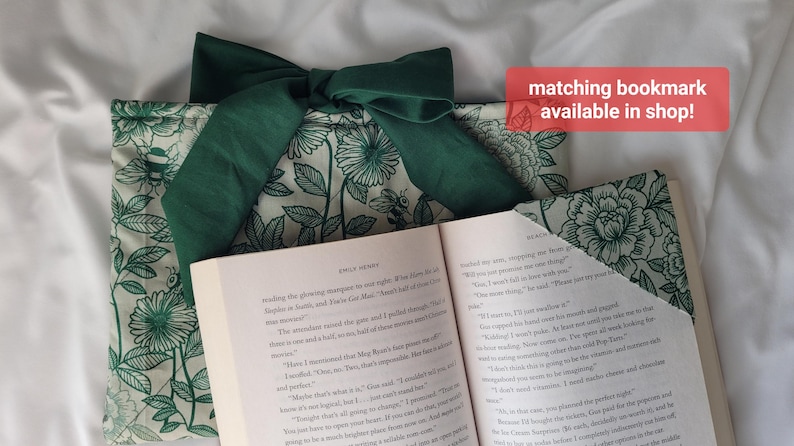 May include: A book sleeve and matching bookmark with a floral pattern in green and white. The book sleeve has a large green bow. The open book displays text. The text "matching bookmark available in shop!" is in a red box.