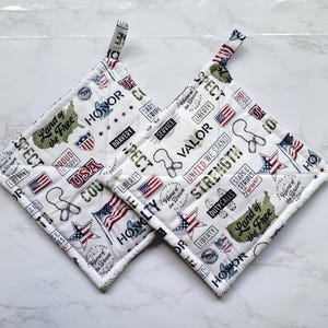 May include: Two white quilted pot holders with patriotic designs. The pot holders feature text and graphics including flags, stars, and phrases like "Land of the Free" and "USA". Each has a loop for hanging.