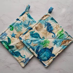 May include: Two square pot holders with a white background and a blue and green sea life pattern. The pattern includes seahorses, starfish, seashells, and a turtle.