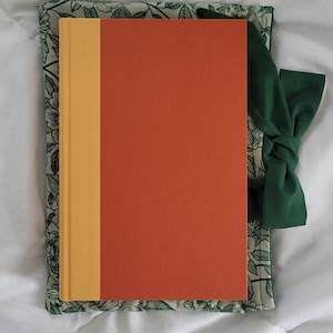 May include: A large book sleeve, 8x11 inches, with a green floral pattern and a dark green bow. An orange and yellow book is inside the sleeve. The text on the image reads "LARGE 8x11in sleeve" and "fits most standard size hardcovers."