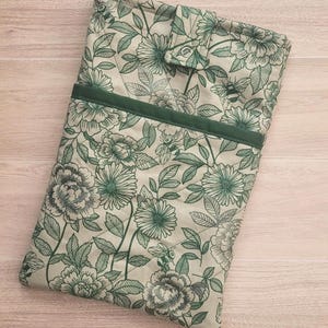 May include: A floral-patterned book sleeve in shades of green and cream. The design features large flowers, leaves, and bees. A dark green band accents the top, and a button closure secures the sleeve. The fabric has a quilted texture.