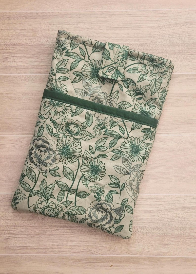 May include: A floral-patterned book sleeve in shades of green and cream. The design features large flowers, leaves, and bees. A dark green band accents the top, and a button closure secures the sleeve. The fabric has a quilted texture.