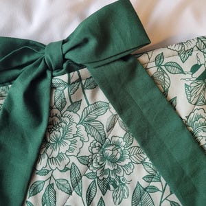 May include: A close-up of a green bow tied on top of a floral patterned fabric. The fabric has a cream background with a green floral design. The bow is a solid, deep green color.