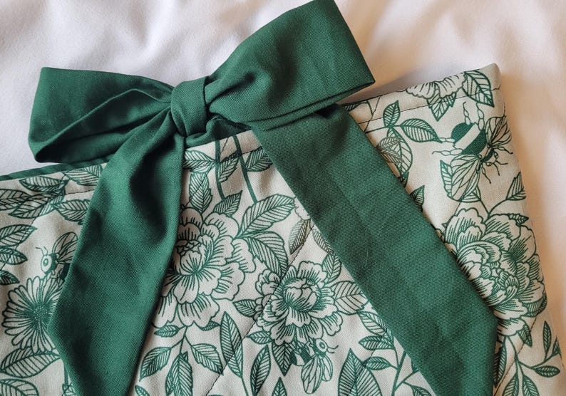 May include: A close-up of a green bow tied on top of a floral patterned fabric. The fabric has a cream background with a green floral design. The bow is a solid, deep green color.