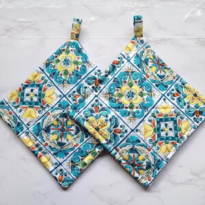 May include: Two square pot holders with a colorful, repeating tile pattern. The design features yellow, teal, and blue floral motifs on a white background. Each pot holder has a small fabric loop for hanging.