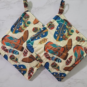 May include: Two square pot holders with a cream background. The pot holders feature a repeating pattern of brown cowboy boots, blue and orange feathers, and blue stars. Each pot holder has a fabric loop for hanging.
