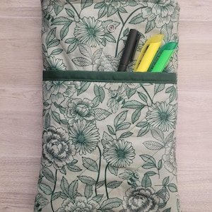 May include: A floral print book sleeve with a snap button closure and front pocket. The sleeve is light green with a pattern of green flowers and leaves. The text "NEW YORK TIMES BESTSELLER" is visible. Includes pens.