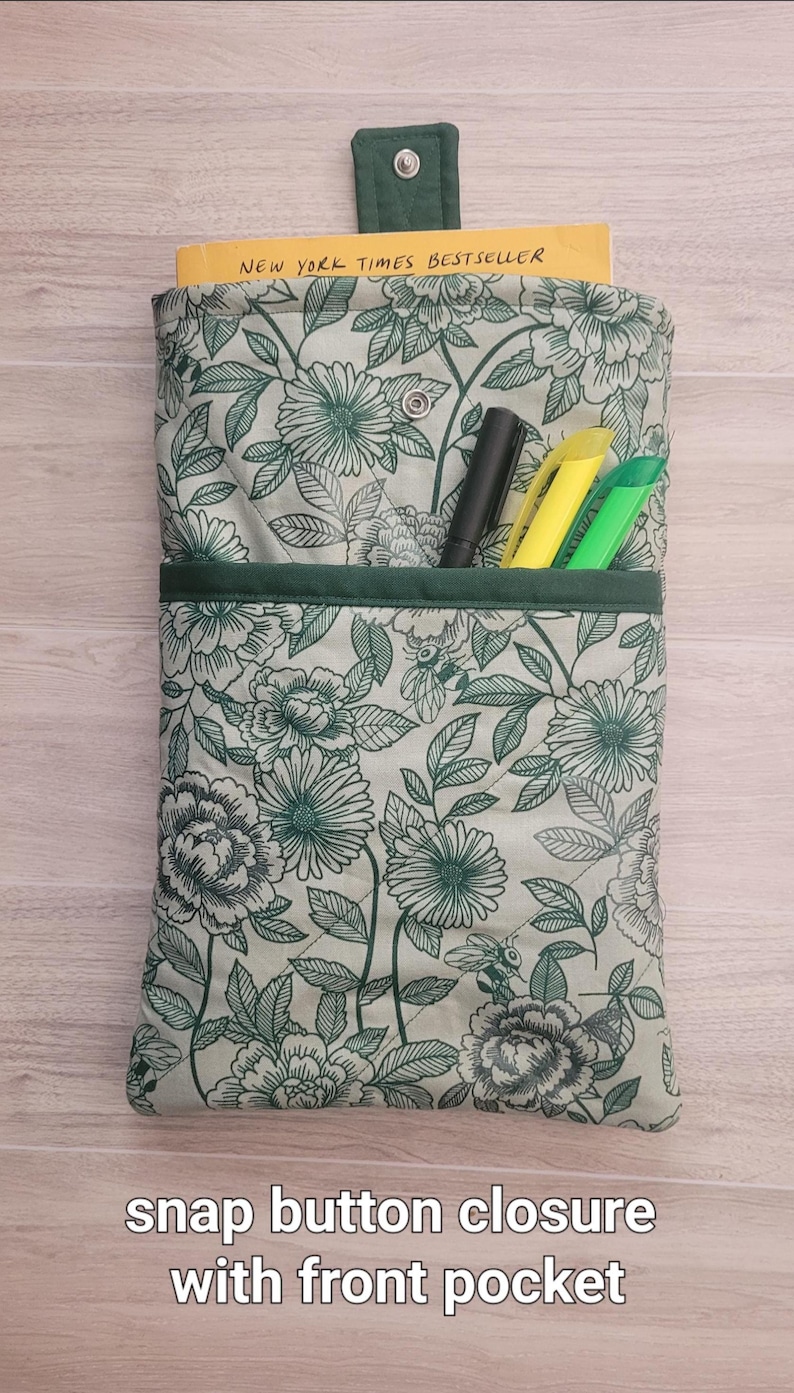 May include: A floral print book sleeve with a snap button closure and front pocket. The sleeve is light green with a pattern of green flowers and leaves. The text "NEW YORK TIMES BESTSELLER" is visible. Includes pens.