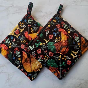 May include: Two square pot holders with a black background featuring a rooster and floral pattern. The roosters are in shades of orange, yellow, and brown. The flowers are red, blue, and yellow. Each pot holder has a black loop for hanging.