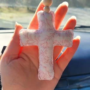 May include: A fabric cross with a pink floral pattern, suspended by a string with wooden beads. The cross is made of soft, padded material and is likely a decorative item or ornament.