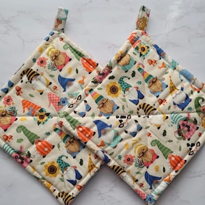 May include: Two white quilted pot holders with a gnome pattern. The gnomes are wearing colorful hats and clothes. The background is a cream color with sunflowers and other floral accents.