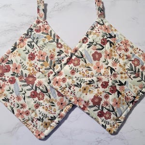 May include: Two quilted pot holders with a floral pattern in shades of pink, red, yellow, and green on a white background. The pot holders have a loop for hanging.