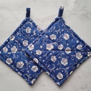May include: Two blue quilted pot holders with a floral pattern of white and gray flowers and vines. Each pot holder has a small fabric loop for hanging. The pot holders are square-shaped.
