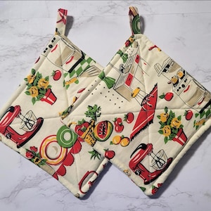 May include: Two square pot holders with a retro kitchen design. The cream-colored fabric features red stand mixers, tomatoes, and yellow flowers. Each pot holder has a loop for hanging.
