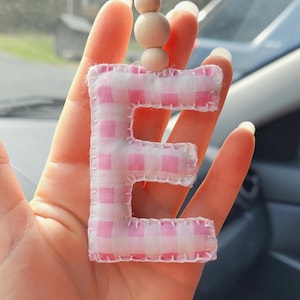 May include: A fabric letter "E" car charm with a pink and white gingham pattern. The charm is stitched with white thread and has a string with wooden beads for hanging. The background is blurred, suggesting it's in a car.