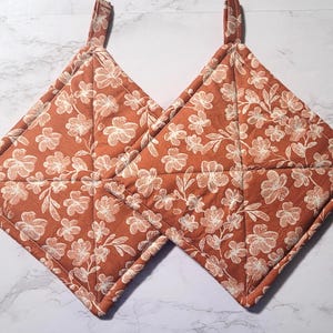 May include: Two rust-colored quilted pot holders with a white floral pattern. The pot holders have a loop for hanging.