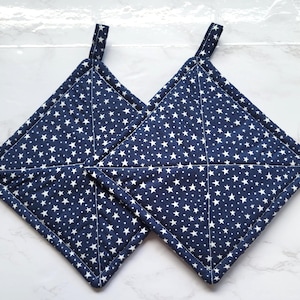May include: Two navy blue, square potholders with white star patterns. Each potholder has a small loop for hanging. The potholders are made of fabric and are designed for kitchen use.