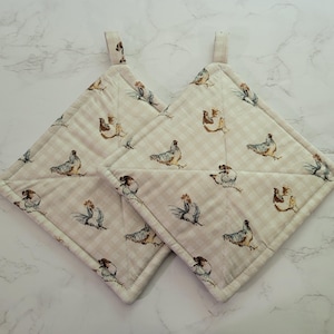 May include: Two square quilted pot holders featuring a light beige and white gingham check pattern. The fabric is decorated with a repeating print of various chickens and roosters in shades of brown, gray, and white. Each pot holder has a fabric loop.