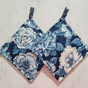 May include: Two square pot holders with a floral pattern. The pot holders feature a navy blue background with large white and light blue flowers. Each pot holder has a small fabric loop for hanging.