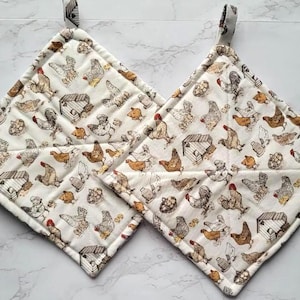 May include: Two quilted, square pot holders with a white background and a repeating pattern of chickens, chicken coops, and eggs in brown, tan, and red. Each pot holder has a hanging loop.