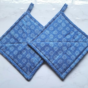 May include: Two blue quilted pot holders with a white circular pattern. Each pot holder is square with a loop for hanging. The edges are trimmed with a darker blue fabric. The pot holders are designed for kitchen use.