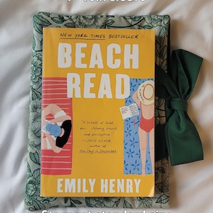 May include: A book sleeve with a floral pattern, holding a paperback book titled "Beach Read" by Emily Henry. The sleeve is approximately 7 x 10 inches and has a dark green bow. The book cover is yellow with illustrations of people on a beach.