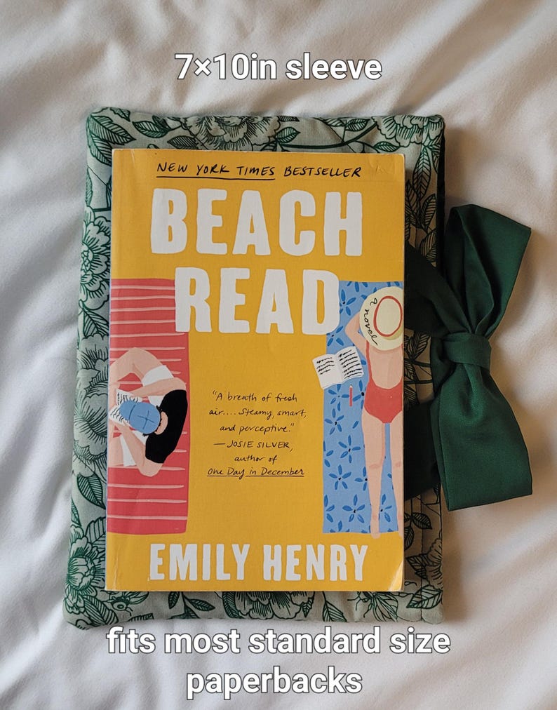 May include: A book sleeve with a floral pattern, holding a paperback book titled "Beach Read" by Emily Henry. The sleeve is approximately 7 x 10 inches and has a dark green bow. The book cover is yellow with illustrations of people on a beach.