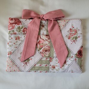May include: A rectangular fabric pouch featuring a patchwork design of floral patterns in shades of pink, green, and white. The pouch is accented with a large, solid dusty pink fabric bow tied at the top center, adding a decorative element.