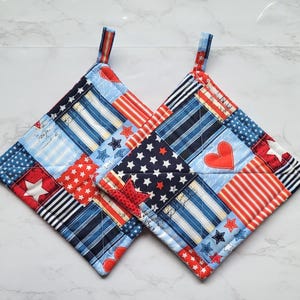 May include: Two square pot holders with patriotic patchwork designs. Each features red, white, and blue fabrics with stars, stripes, and heart shapes. They have small fabric loops for hanging.