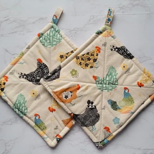 May include: Two beige quilted pot holders with a pattern of colorful chickens and flowers. The pot holders have a loop for hanging.