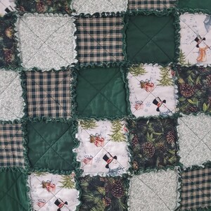 May include: A patchwork quilt with a festive winter theme. The quilt features squares of green, white, and plaid fabrics. Some squares display snowmen, pinecones, and Christmas trees. The edges are finished with a ruffled trim, creating a cozy, handmade aesthetic.