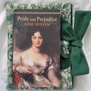 May include: A small, green floral book sleeve, 6x8 inches, with a copy of "Pride and Prejudice" by Jane Austen. The book is nestled inside the sleeve, with a matching green tie. The text "fits most pocket size books" is also visible.