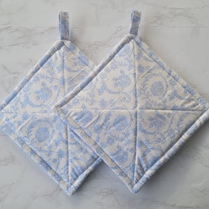 May include: Two square quilted pot holders with a floral pattern in light blue and cream. Each pot holder has a loop for hanging. The pot holders are approximately 8 inches square.