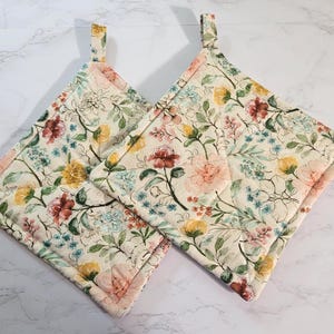 May include: Two quilted pot holders with a floral pattern. The fabric features a cream background with a repeating design of pink, yellow, and burgundy flowers, along with green leaves and blue accents. Each pot holder has a small fabric loop for hanging.