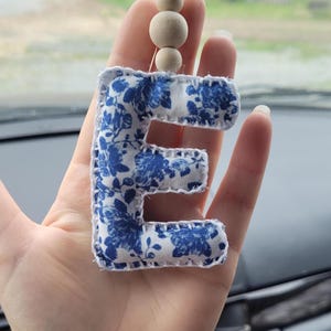 May include: A fabric letter "E" car charm with a blue floral pattern on a white background. The charm is stitched with white thread and has wooden beads attached. The charm is hanging in a car.