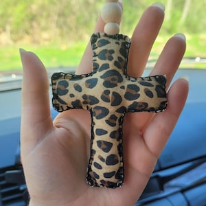 May include: A decorative cross-shaped car charm. The charm features a leopard print pattern in shades of brown and black. It is trimmed with black stitching and has wooden beads and a black string for hanging.