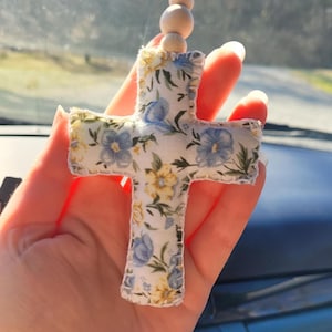 May include: A floral cross-shaped car charm. The charm is made of fabric with a pattern of blue and yellow flowers on a white background. It is suspended by a string with wooden beads. The cross is held in a hand.