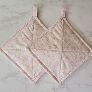 May include: Two quilted pot holders with a pink floral pattern on a white background. Each square-shaped holder has a loop for hanging and is stitched with a diamond pattern. The edges are trimmed with a pink binding.