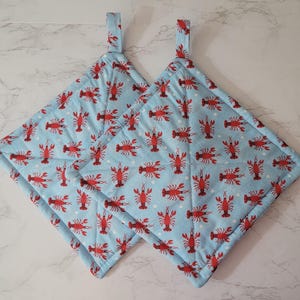 May include: Two square quilted pot holders featuring a light blue background with a repeating pattern of small red lobsters and white dots. Each pot holder has a fabric loop at the top corner for hanging, with stitched diagonal lines.