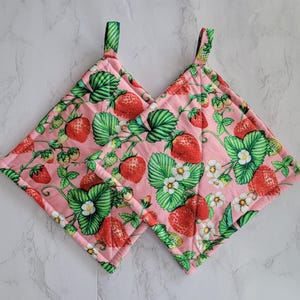 May include: Two pink, square pot holders with a strawberry pattern. The design features red strawberries, green leaves, and white flowers on a pink background. Each pot holder has a green fabric loop for hanging.