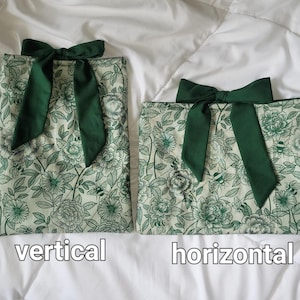 Peut inclure: Deux pochettes en tissu avec un motif floral et d'abeilles vert sur fond cr&egrave;me. Chaque pochette est orn&eacute;e d'un n&oelig;ud en tissu vert fonc&eacute; sur le dessus. La pochette de gauche est orient&eacute;e verticalement et celle de droite horizontalement.