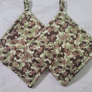 May include: Two square pot holders with hanging loops. The pot holders feature a camouflage pattern in shades of green, brown, and tan. The edges are neatly sewn, and the fabric appears to be quilted.