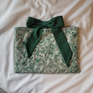 May include: A rectangular, floral-patterned pouch with a dark green bow. The pouch features a cream background with a green floral design. The bow is made of a solid dark green fabric. The pouch is likely for storing items.
