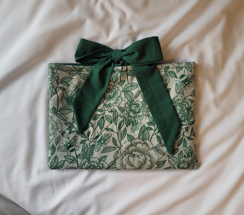 May include: A rectangular, floral-patterned pouch with a dark green bow. The pouch features a cream background with a green floral design. The bow is made of a solid dark green fabric. The pouch is likely for storing items.