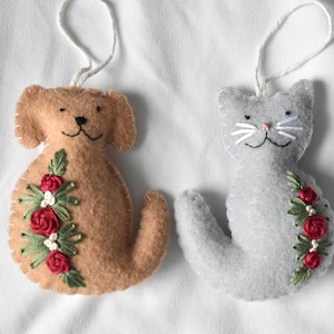 May include: Two felt ornaments shaped like a tan dog and a gray cat, each featuring embroidered red roses and green leaves on the side, stitched edges, and a white string loop at the top for hanging, crafted from soft felt material.