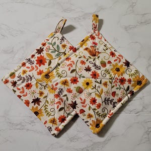 May include: Two square quilted pot holders featuring a cream-colored fabric with a repeating autumn-themed pattern of yellow sunflowers, orange flowers, brown maple leaves, green vines, small red mushrooms, acorns, and a small brown squirrel.