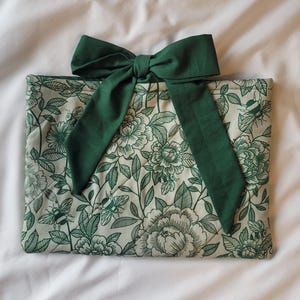May include: A floral-patterned clutch with a dark green bow. The clutch features a cream background with a green floral design. The bow is made of a matching dark green fabric. The clutch is lying on a white surface.