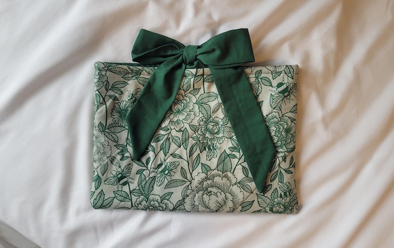 May include: A floral-patterned clutch with a dark green bow. The clutch features a cream background with a green floral design. The bow is made of a matching dark green fabric. The clutch is lying on a white surface.