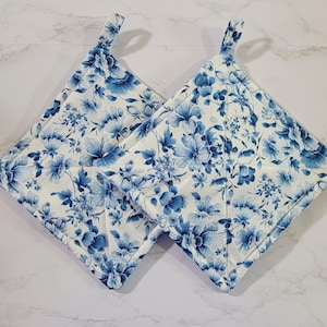 May include: Two square pot holders with a white background and a blue floral pattern. Each pot holder has a loop for hanging. The floral design features various blue flowers and leaves.